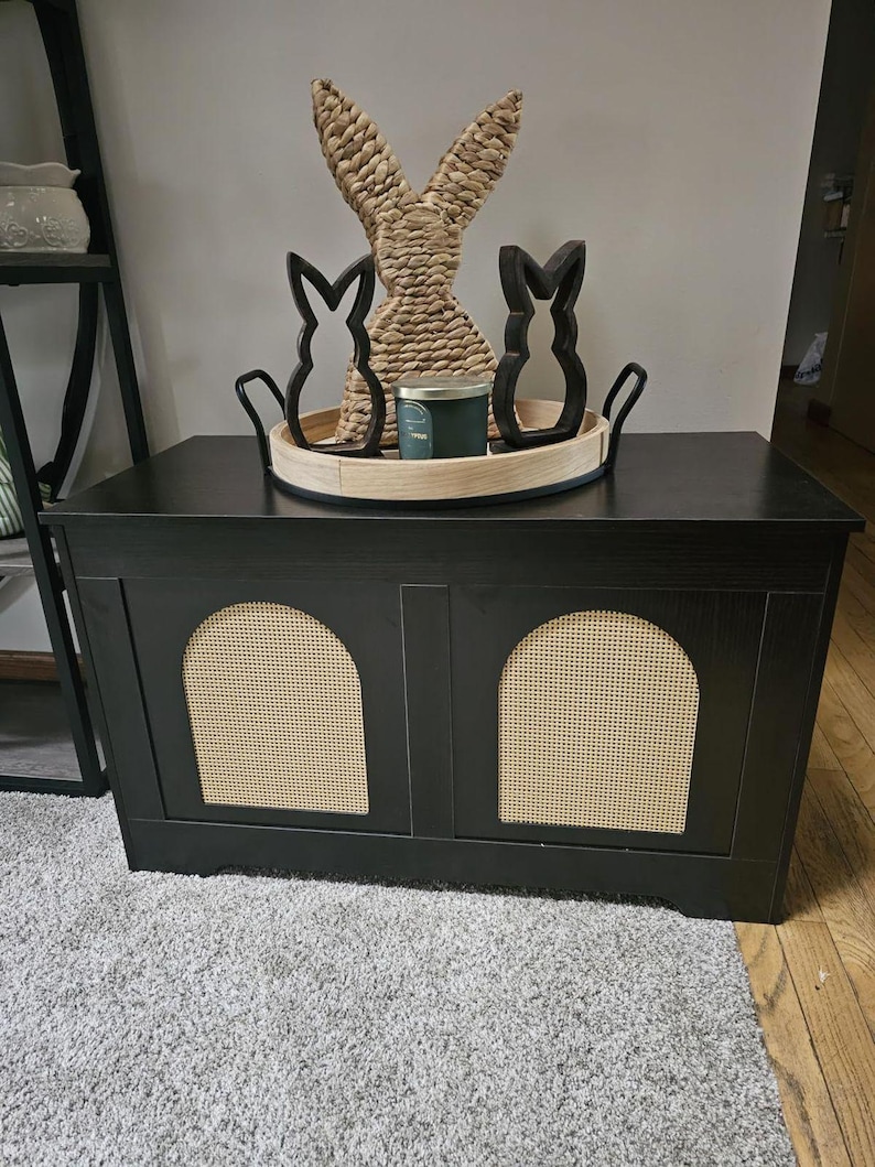 May include: A black storage cabinet with two arched rattan panels. A wooden tray on top holds a woven rabbit figure, two black bunny silhouettes, and a green candle. The cabinet sits on a grey carpet.