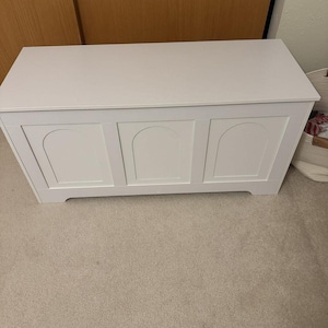 May include: A white wooden storage chest with a flat top and three arched panels on the front. The chest is painted white and sits on a beige carpeted floor. The chest is designed for storing items, a practical storage solution.