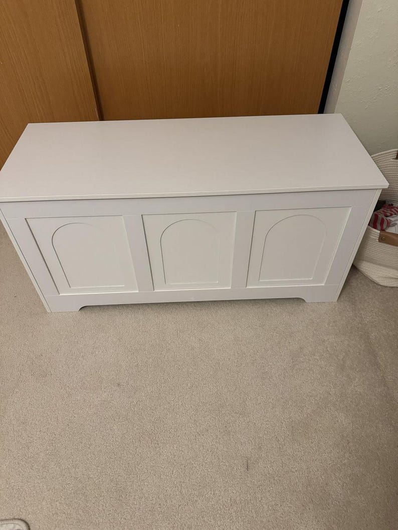 May include: A white wooden storage chest with a flat top and three arched panels on the front. The chest is painted white and sits on a beige carpeted floor. The chest is designed for storing items, a practical storage solution.