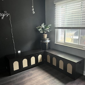 May include: Two matching black storage benches with arched, beige panels. The benches are in a corner of a room with dark grey walls and a dark wood-effect floor. A plant sits on a decorative stand.