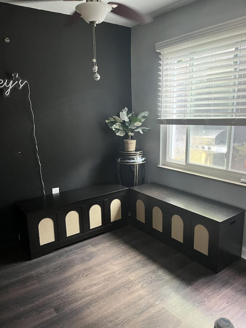 May include: Two matching black storage benches with arched, beige panels. The benches are in a corner of a room with dark grey walls and a dark wood-effect floor. A plant sits on a decorative stand.