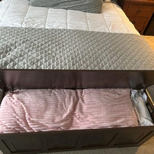 May include: A dark brown wooden storage chest with the lid partially open, revealing a pink blanket and a white pillow inside. The chest is placed at the foot of a bed with a grey quilted bedspread.