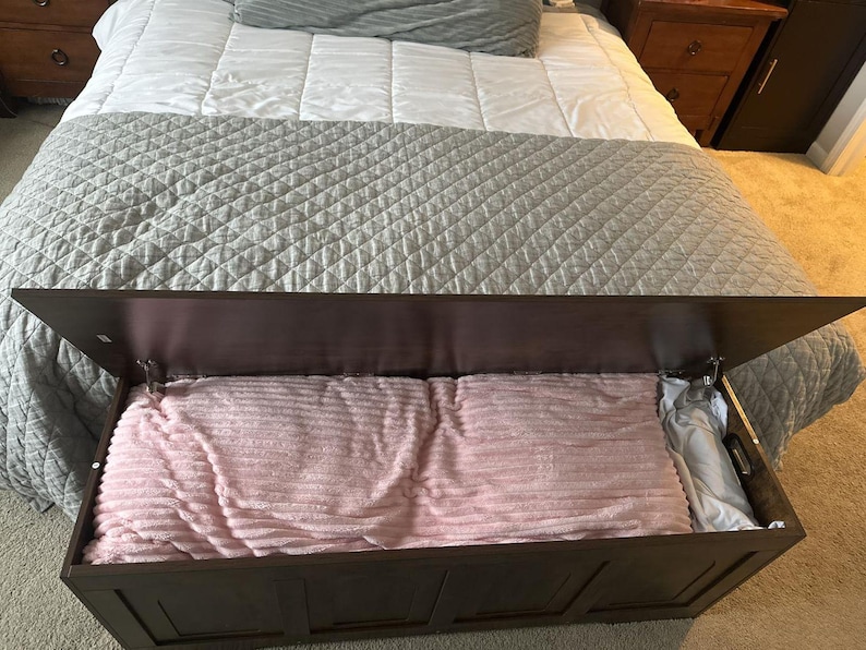 May include: A dark brown wooden storage chest with the lid partially open, revealing a pink blanket and a white pillow inside. The chest is placed at the foot of a bed with a grey quilted bedspread.