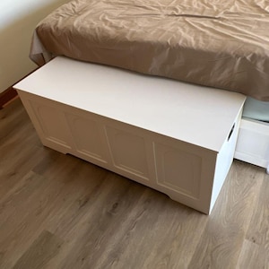 May include: A white wooden storage chest with a rectangular shape and decorative panels. The chest is positioned at the foot of a bed with a brown duvet. The chest is on a wooden floor.