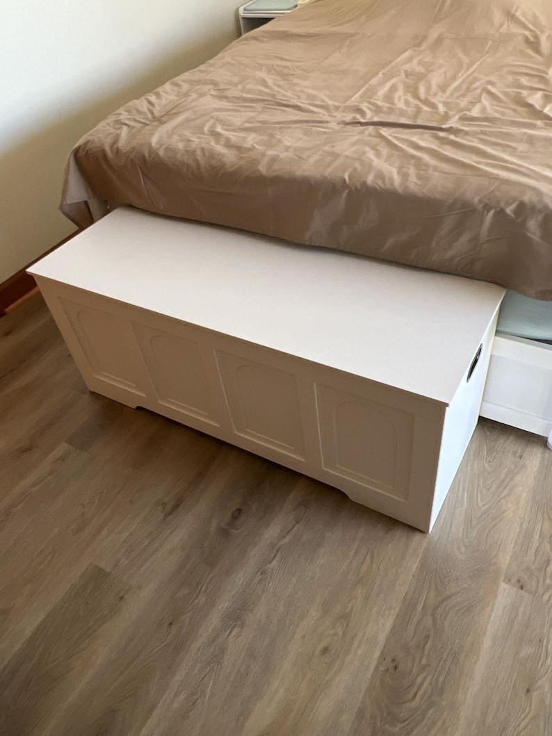 May include: A white wooden storage chest with a rectangular shape and decorative panels. The chest is positioned at the foot of a bed with a brown duvet. The chest is on a wooden floor.