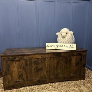 May include: A dark brown wooden storage chest with arched panels. A fluffy white sheep toy and a sign saying "Love you more" are on top. The chest sits on a woven rug against a blue wall.
