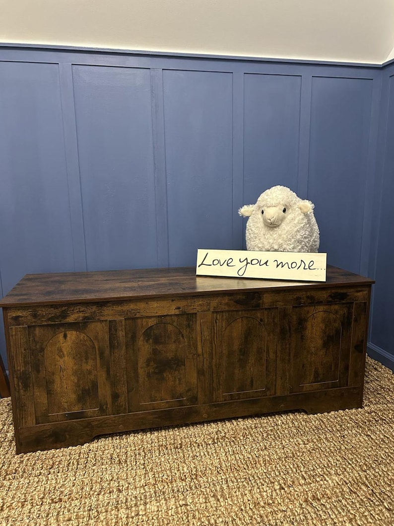 May include: A dark brown wooden storage chest with arched panels. A fluffy white sheep toy and a sign saying "Love you more" are on top. The chest sits on a woven rug against a blue wall.