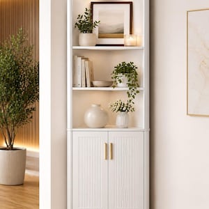 May include: A white corner bookshelf with a cabinet base and open shelves. The shelves display books, plants in pots, a framed landscape picture, and decorative vases. The cabinet doors have vertical grooves and gold-colored handles. The bookshelf is in a well-lit room.