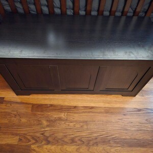 May include: A dark brown wooden storage chest with a flat top and three recessed panels on the front. The chest is positioned on a hardwood floor, with a cot visible in the background.