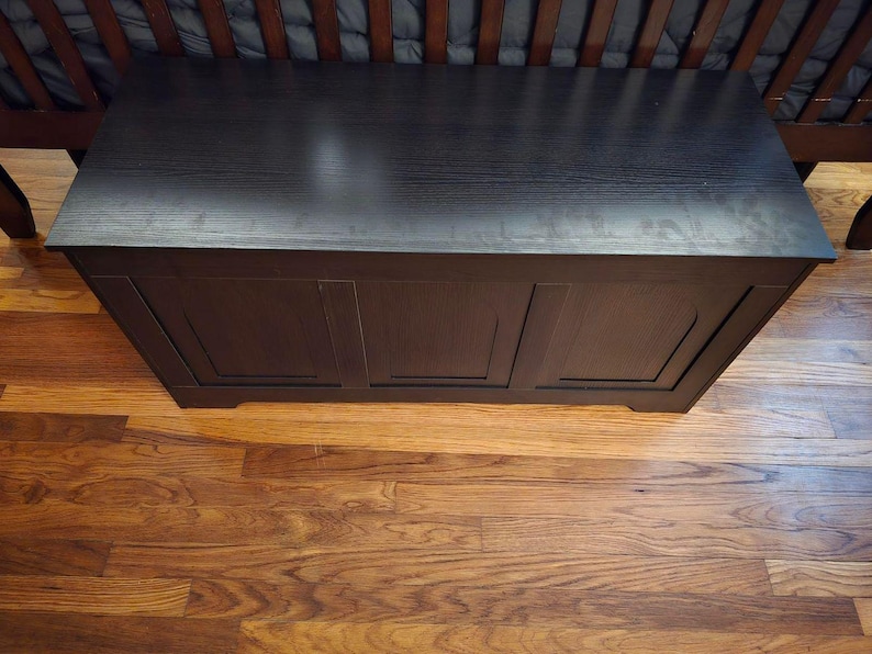 May include: A dark brown wooden storage chest with a flat top and three recessed panels on the front. The chest is positioned on a hardwood floor, with a cot visible in the background.