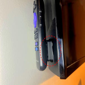 May include: A black Roku TV remote control is held in place by a black plastic clip attached to the side of a black television. The remote has purple and blue buttons.