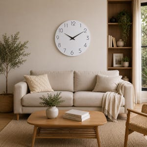 May include: A large circular wall clock with a white face and black numerals from one to twelve, featuring thin black hour and minute hands. The clock is mounted on a plain beige wall above a beige sofa with two throw pillows and a draped blanket.