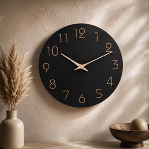 Puede incluir: Un reloj de pared circular con una esfera negra mate y números dorados del uno al doce. El reloj cuenta con dos manecillas delgadas de color dorado situadas en el centro. El diseño es sencillo, con líneas limpias y un esquema de colores contrastado.