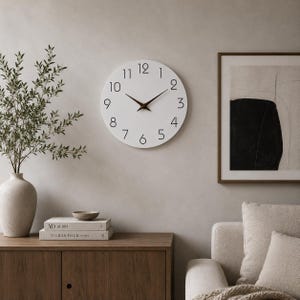 Può includere: Un orologio da parete rotondo con quadrante bianco, numeri neri da 1 a 12 e lancette marrone scuro. L'orologio è montato su una parete liscia sopra un mobile in legno, tra un alto vaso in ceramica con vegetazione e un quadro astratto incorniciato.