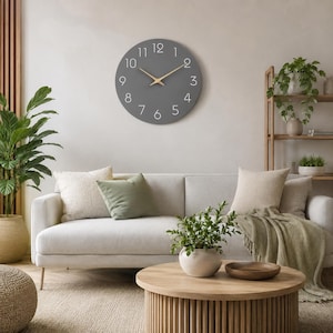 Op de afbeelding: Een ronde wandklok met een mat donkergrijze wijzerplaat, voorzien van witte Arabische cijfers van één tot twaalf en dunne, lichtgekleurde houten wijzers. De klok hangt aan een effen muur boven een lichtgekleurde bank in een woonkamer.