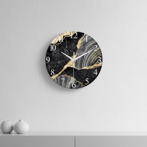 Könnte beinhalten: Runde Wanduhr mit schwarzem Marmormuster und goldenen Akzenten. Das Zifferblatt hat weiße Ziffern und Zeiger. Die Uhr ist an einer weißen Wand befestigt. Die Uhr hat einen Durchmesser von etwa 30 cm.