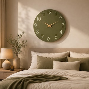 May include: A circular wall clock with a matte olive green face, featuring thin white numerals from one to twelve and two slender, pointed gold-toned hands. The clock is mounted on a plain wall above a bed with pillows and a blanket.