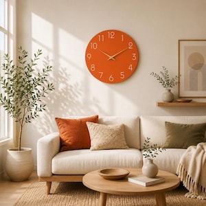 Könnte beinhalten: Eine runde Wanduhr mit einem leuchtend orangefarbenen Zifferblatt, dünnen weißen Ziffern von 1 bis 12 und schlanken goldfarbenen Zeigern. Die Uhr ist an einer schlichten Wand über einem hellen Sofa montiert und dient als zentrales Dekorationselement.