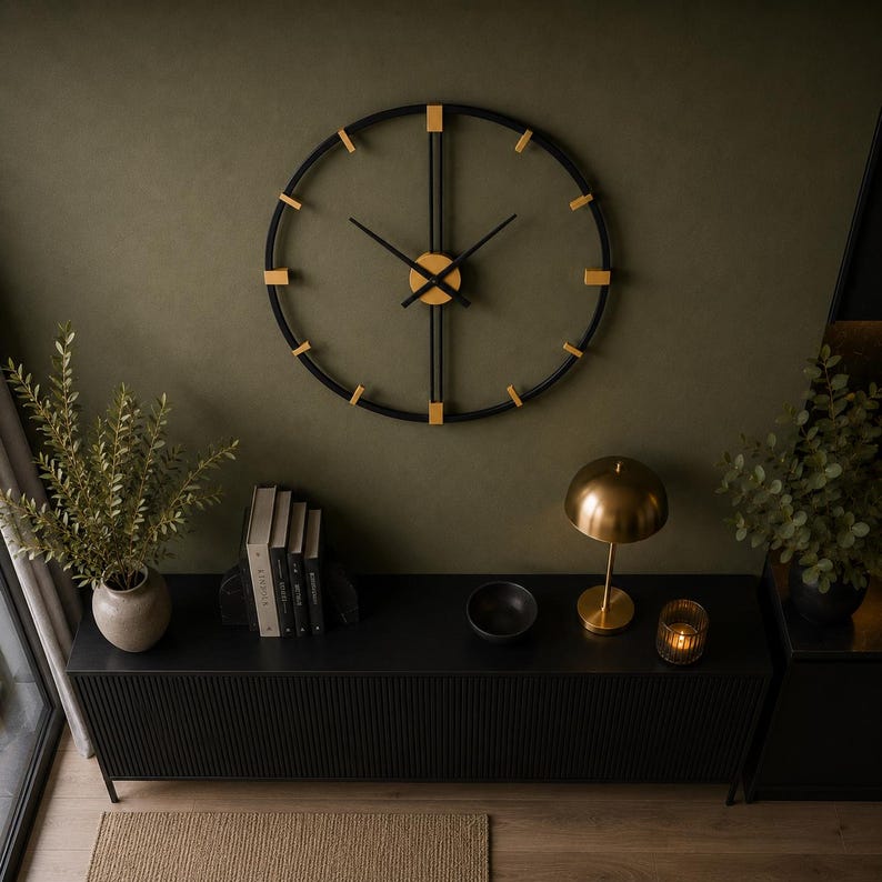 May include: A large circular wall clock featuring a black metal frame with gold-toned accents at each hour marker. The clock has thin black hands and a central gold-toned hub, mounted on a dark olive wall above a black slatted console table.