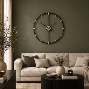 Puede incluir: Un gran reloj de pared circular con un marco fino de metal negro, detalles dorados en los marcadores de hora y un eje central dorado. El reloj cuenta con dos manecillas negras delgadas y una barra de soporte vertical central.