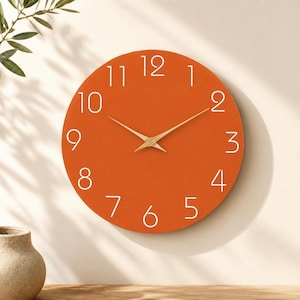 Può includere: Un orologio da parete rotondo con un quadrante color arancione bruciato, caratterizzato da sottili numeri bianchi senza grazie da uno a dodici. L'orologio presenta due lancette in metallo dorato sottili e appuntite al centro del quadrante.