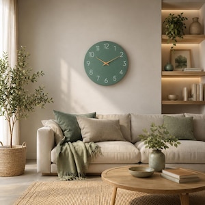 May include: A round wall clock with a matte sage green face, featuring white numerical markers from 1 to 12 and thin, light-colored wooden clock hands. The clock is mounted on a plain, neutral-toned wall above a beige sofa with various cushions.