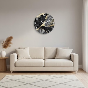 Puede incluir: Un reloj de pared circular con un patrón similar al mármol en negro y gris con vetas de color dorado. La esfera muestra números arábigos blancos del uno al doce e incluye manecillas blancas delgadas para horas y minutos.