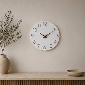 Pode incluir: Um relógio de parede circular com mostrador branco e algarismos pretos de um a doze. O relógio apresenta dois ponteiros pontiagudos castanho-escuros no centro. O design é simples e limpo, exibindo as horas claramente com algarismos arábicos.