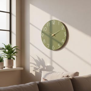 Peut inclure: Une horloge murale ronde vert olive avec des chiffres blancs et des aiguilles dorées. L'horloge est fixée sur un mur clair. Le design est minimaliste et moderne. Le diamètre de l'horloge est d'environ 30 cm. Idéale pour la décoration intérieure.