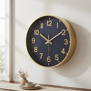 Könnte beinhalten: Eine runde Wanduhr mit einem dunkel marineblauen Zifferblatt, goldenen arabischen Ziffern sowie passenden goldenen Stunden-, Minuten- und Sekundenzeigern. Die Uhr ist in einem glatten, kreisförmigen goldenen Rahmen mit schlichtem Design eingefasst.