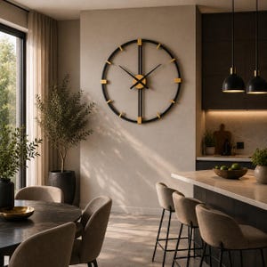 May include: A large circular wall clock featuring a black metal frame with gold-colored accents at the hour markers. The clock has thin black hands and a central gold-colored circular hub, mounted on a plain wall above a kitchen counter area.