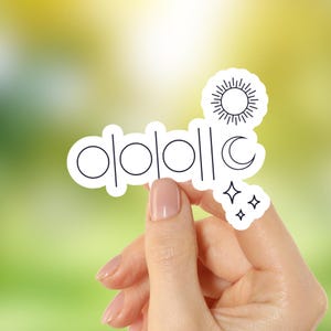 Puede incluir: Una pegatina blanca con un dibujo lineal negro de elementos celestes, incluyendo el sol, la luna y las estrellas. La pegatina también presenta una serie de círculos y líneas verticales. La pegatina está sujeta por una persona.