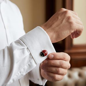Peut inclure: Gros plan sur un poignet de chemise blanche avec un bouton de manchette en forme de coccinelle. Le bouton de manchette est rouge avec des détails noirs et deux petits points noirs. La chemise est en tissu blanc.
