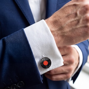 May include: Silver cufflinks with a black vinyl record design and a red center label. The cufflinks are on a white dress shirt cuff, paired with a navy blue suit jacket. The image is a close-up shot.