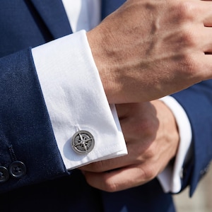 Puede incluir: Primer plano de gemelos plateados con forma de brújula en un puño de camisa blanca, combinados con un traje azul marino. Los gemelos presentan un diseño detallado de rosa de los vientos. La imagen resalta el estilo clásico y la artesanía.