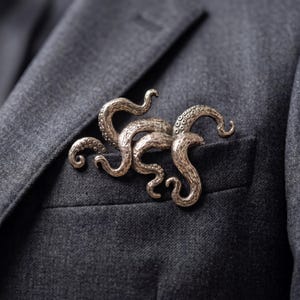 Puede incluir: Broche plateado con forma de pulpo y tentáculos detallados, prendido a una chaqueta gris. La broche tiene una superficie texturizada y está colocada cerca del bolsillo de la chaqueta.
