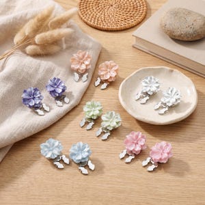 Peut inclure: Diverses barrettes en forme de fleurs aux couleurs pastel, rose, pêche, lavande, bleu clair, vert et blanc. Chaque barrette est dotée d'une base en métal argenté. Les barrettes sont disposées sur une surface en bois.