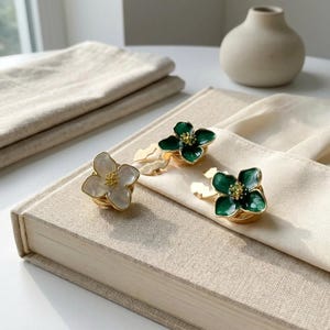 Op de afbeelding: Drie bloemvormige servetringen. Eén is wit met een gouden centrum en twee zijn groen met gouden centra. De servetringen liggen op een beige boek en een beige servet. Een kleine beige vaas staat op de achtergrond.