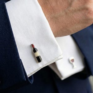 Może przedstawiać: Spinki do mankietów z miniaturowym wzorem butelki wina z czerwoną, czarną i białą emalią. Spinki do mankietów są umieszczone na białym mankiecie koszuli i granatowym garniturze.