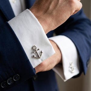 Può includere: Gemelli a forma di ancora argentata su un polsino di camicia bianca, abbinati a una giacca blu navy. I gemelli sono dettagliati e lucenti. L'immagine si concentra sui dettagli dell'abbigliamento formale.