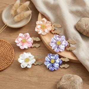 Op de afbeelding: Vijf bloemvormige servetringen in tinten roze, perzik, wit, paars en blauw met goudkleurige accenten. De bloemmotieven hebben een textuur en zijn gerangschikt op een houten oppervlak.