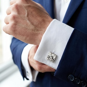 Puede incluir: Gemelos cuadrados plateados con un diseño de cordillera. Los gemelos se lucen en un puño de camisa blanca, combinados con una chaqueta de traje azul marino. El diseño de la montaña es detallado y texturizado.
