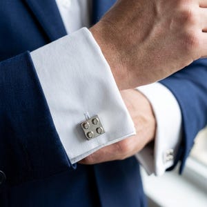 May include: Silver cufflinks with a square design featuring four circular accents. The cufflinks are attached to a white dress shirt cuff, complemented by a navy blue suit jacket. The image is a close-up shot.
