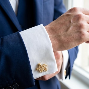 Peut inclure: Boutons de manchette dorés en forme de serpent sur une manchette de chemise blanche. Les boutons de manchette représentent un serpent enroulé. La chemise est portée avec une veste de costume bleu marine.