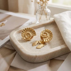 May include: A pair of gold-coloured earrings featuring a spiral seashell design. One is a stud, the other has a dangling element. The earrings are presented on a light-coloured tray, with a neutral background.