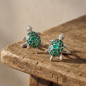 May include: A pair of silver cufflinks shaped like turtles. Each turtle has a green, textured shell and detailed flippers and head. The cufflinks are displayed on a rustic wooden surface.