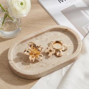 May include: Two gold-coloured, flower-shaped candle holders sit on a beige, oval marble tray. One holder is solid, while the other has a circular indentation for a candle. A small glass vase with a white flower is in the background.