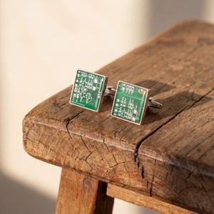 May include: Square cufflinks featuring a green circuit board design, set in a silver frame. The cufflinks are displayed on a rustic wooden surface, highlighting their intricate details and modern aesthetic. The cufflinks are approximately 1.5 cm square.