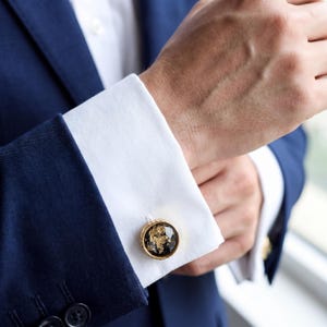 Puede incluir: Primer plano de un traje azul marino con camisa blanca y gemelos dorados. Los gemelos tienen un fondo negro con detalles de pan de oro y un borde dorado. La chaqueta del traje tiene botones negros.
