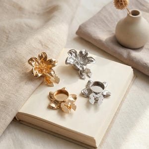 May include: Four flower-shaped candle holders in gold and silver. The candle holders are on an open book and a beige cloth. A small vase with a dried flower is in the background.